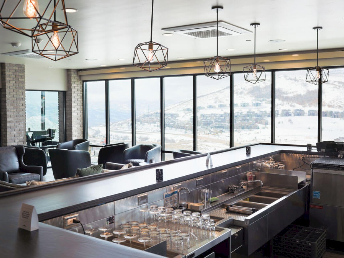 A modern indoor bar area with large windows overlooking a scenic view. The bar features glasses, sinks, and contemporary light fixtures.