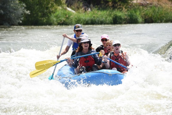 A group of people is white-water rafting in a river, wearing life jackets and helmets, with smiles and excitement on their faces.