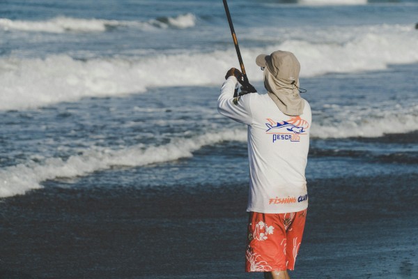 A person wearing a hat and white shirt is fishing at the beach, facing the ocean waves, holding a fishing rod.