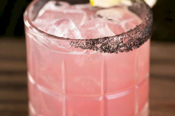 This image shows a pink cocktail served over ice in a glass with a black salt rim and a slice of lime on the rim.