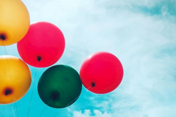 The image shows five balloons of various colors floating against a bright blue sky with some clouds in the background.