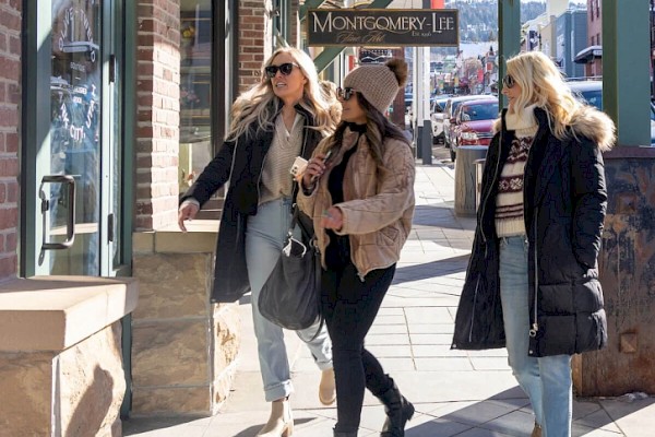 Three women dressed in winter clothing walk along a sidewalk, with a store named 