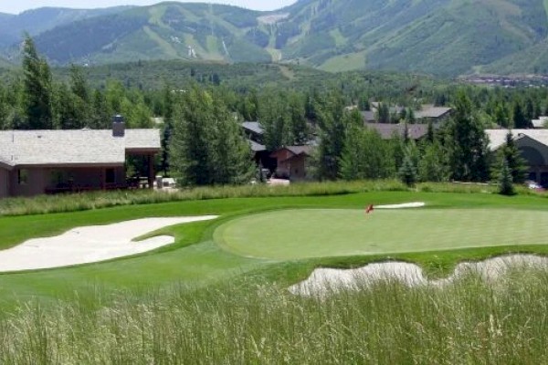 A golf course with sand traps, a green flagstick, houses in the background, and mountainous terrain in the distance.