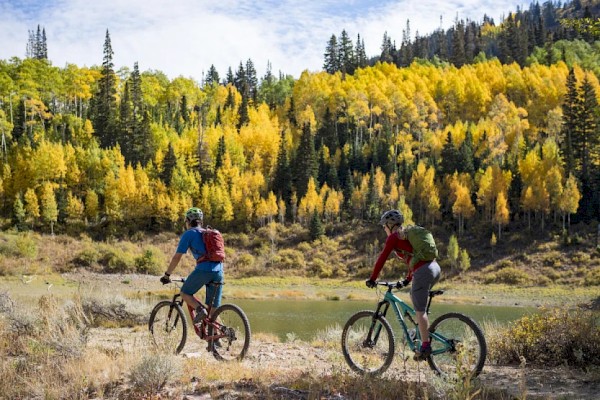 Two people riding mountain bikes on a trail with a scenic background of autumnal trees and hills.