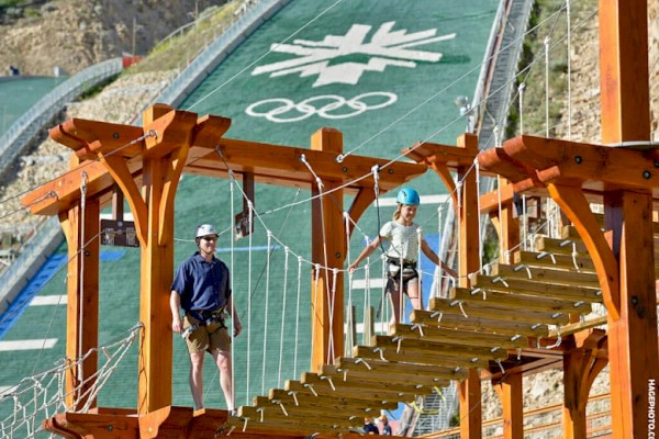 Two people wearing helmets navigate a wood and rope bridge with an Olympic ski jump in the background, featuring the Olympic rings and a snowflake design.