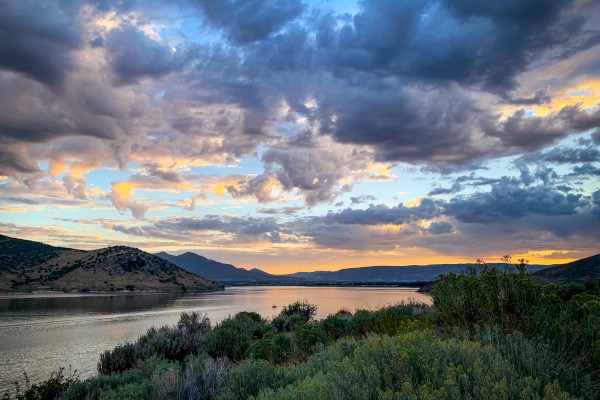 A serene river landscape at sunset with mountains, partly cloudy sky, and lush green vegetation in the foreground.