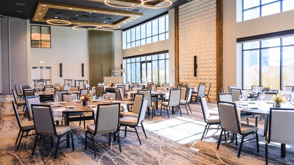 A spacious banquet hall with round tables and cushioned chairs, set for a formal event, large windows and modern ceiling lights.