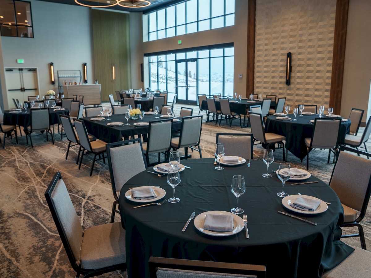 A modern banquet hall set up with round tables, each adorned with plates, silverware, and wine glasses. Large windows on the side.