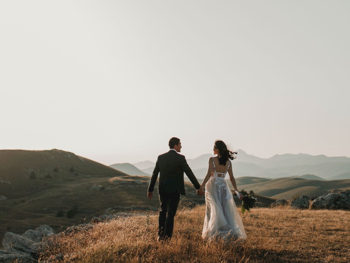 A couple dressed in wedding attire walks hand in hand across a scenic, grassy landscape with rolling hills in the background.