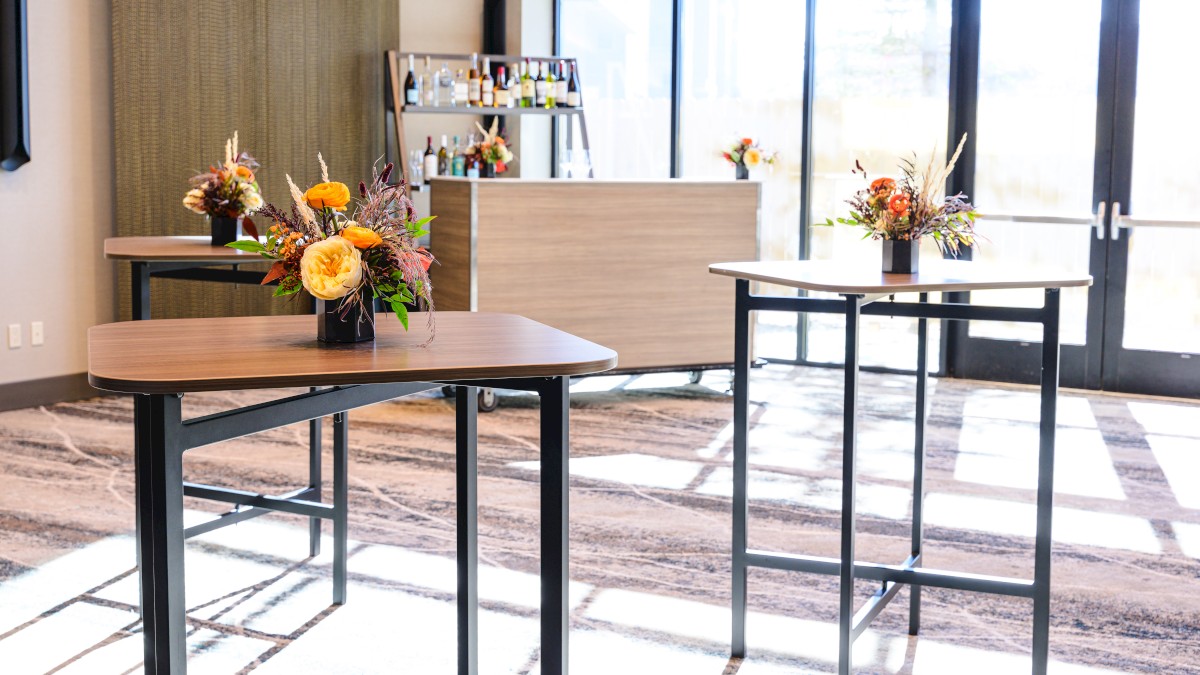 A bright lobby with two tall round tables, flower arrangements, and a reception area with a shelf of bottles in the background, near large glass doors.