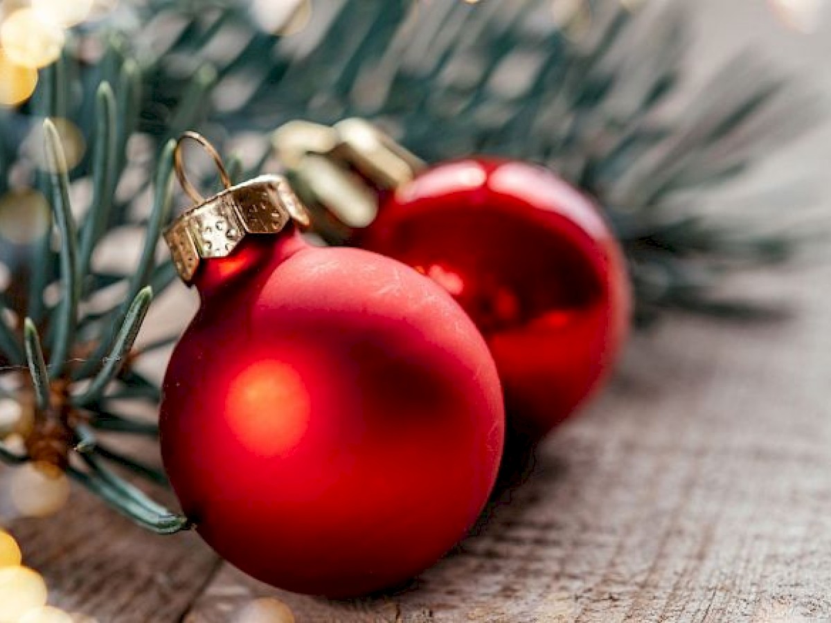 The image shows red Christmas ornaments on a wooden surface next to some pine branches with holiday lights blurred in the background.