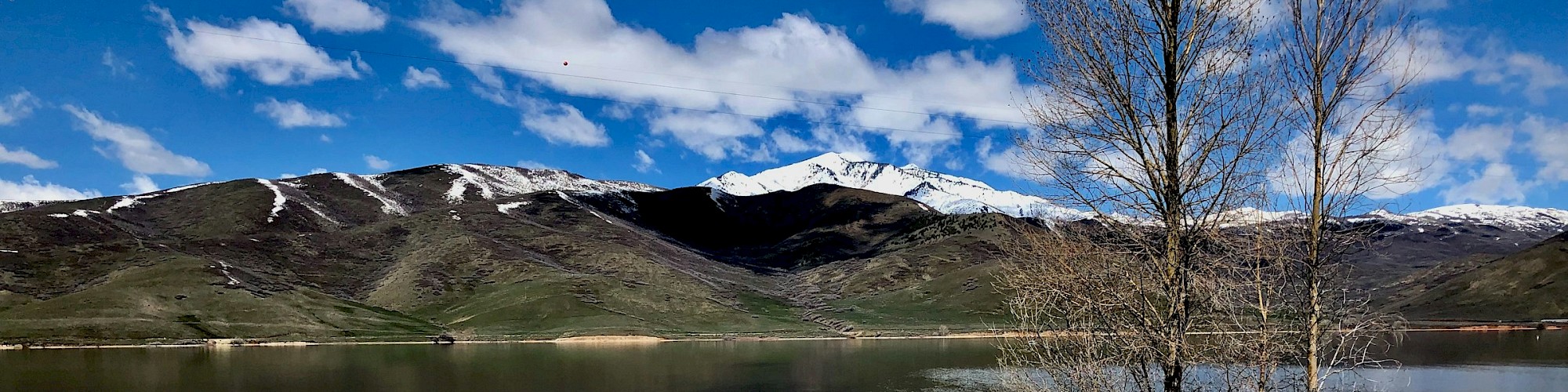 A serene lake with reflections, surrounded by snow-capped mountains, under a bright blue sky with scattered clouds.