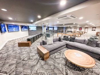 The image shows a modern lounge area with multiple TVs, grey couches, patterned carpet, wooden tables, and bright, ambient lighting.