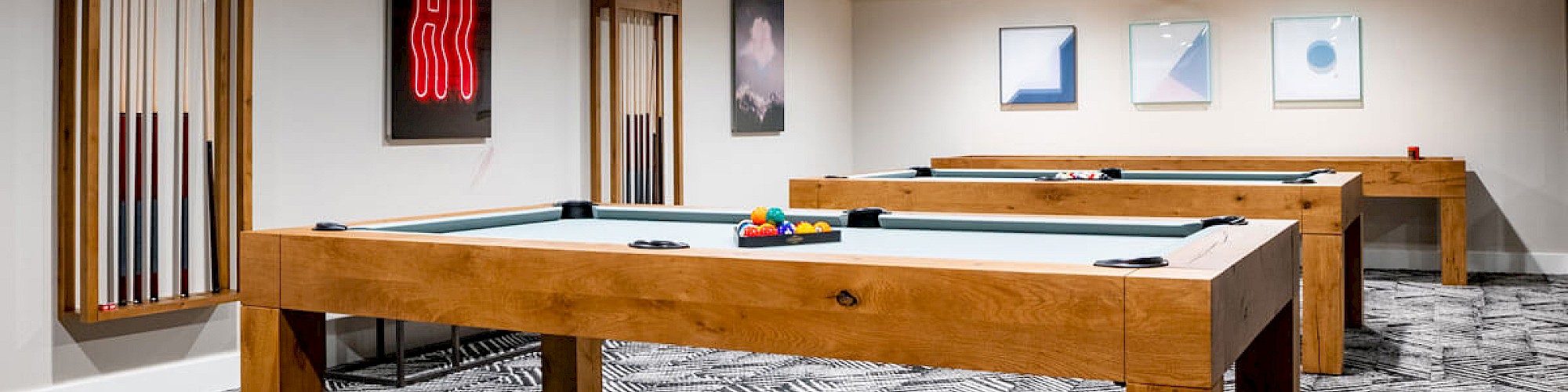 The image shows a modern room with multiple pool tables, framed artwork on the walls, and a patterned carpet.