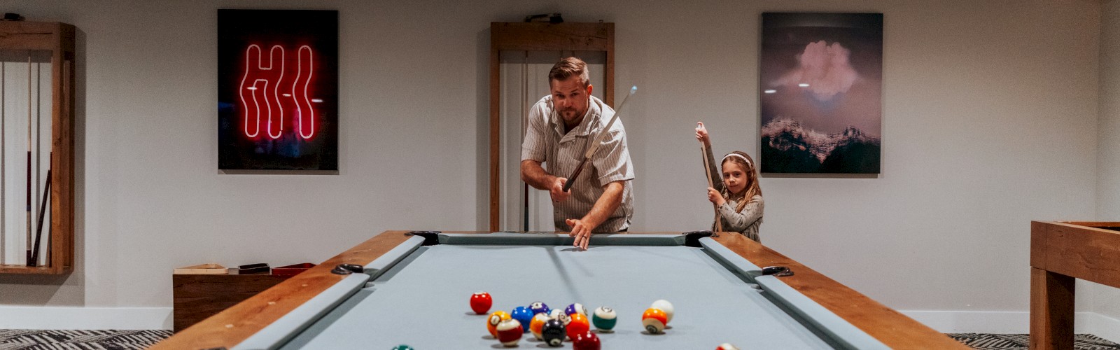 A man and child play pool in a room with neon art and photo on the walls.
