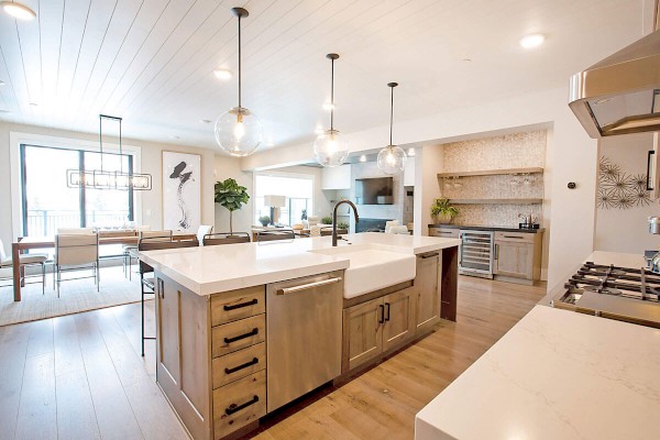 A modern kitchen with a large island, pendant lights, and wooden cabinets opens to a dining area with a table and chairs.