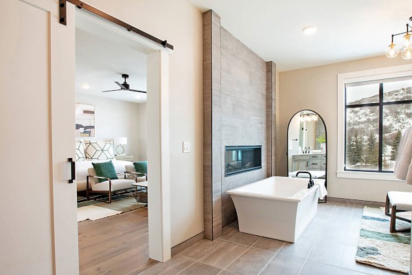 A modern bathroom with a white bathtub, a fireplace, an arch mirror, a window, and a sliding door leading to a living room.