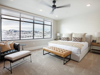 The image shows a modern bedroom with a large window view of snowy mountains, a bed with cushions, a bench at the foot, and a sitting area.