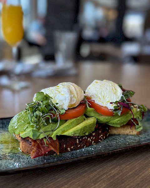 Open-faced avocado toast with poached egg, tomato, greens, and crispy bread on a stylish plate, served in a cozy restaurant setting.