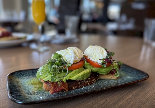 Open-faced avocado toast with poached egg, tomato, greens, and crispy bread on a stylish plate, served in a cozy restaurant setting.