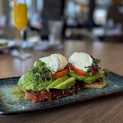 Open-faced avocado toast with poached egg, tomato, greens, and crispy bread on a stylish plate, served in a cozy restaurant setting.