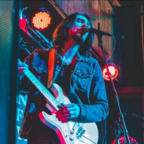 A musician plays an electric guitar and sings into a microphone under colorful stage lights, wearing a denim jacket and a necklace with a cross pendant.