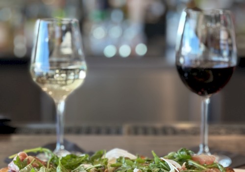 A pizza topped with greens and onions is in the foreground, with glasses of white and red wine placed behind it on a table, in a restaurant.