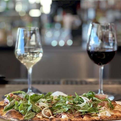 A pizza topped with greens and onions is in the foreground, with glasses of white and red wine placed behind it on a table, in a restaurant.