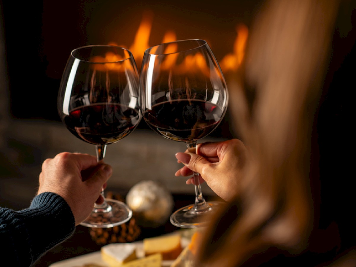 Two people clinking glasses of red wine near a fire. A cozy atmosphere with cheese and snacks visible in the background.