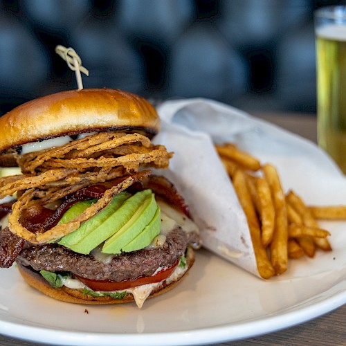 A burger with avocado, onion rings, fries, and a drink on a white plate. The background is blurred, emphasizing the food in focus.