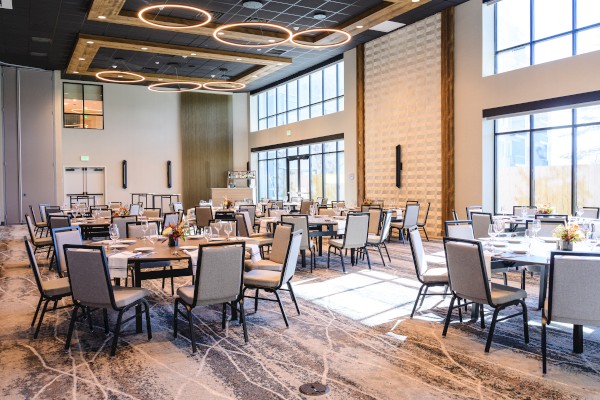 A spacious, modern event hall with round and square tables, grey chairs, large windows, and stylish ceiling lights.