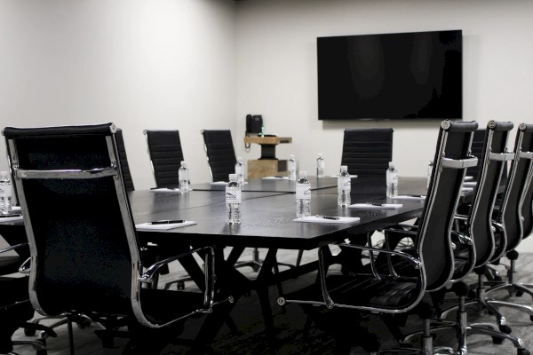 A modern conference room with a long table, black chairs, bottled water, notepads, and a large wall-mounted TV.