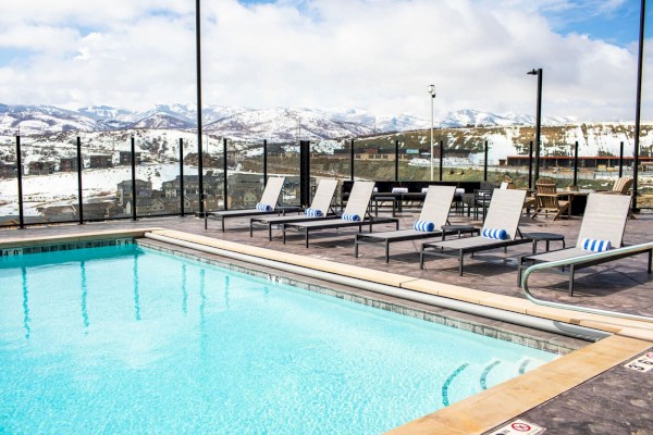 An outdoor swimming pool with several lounge chairs lined up beside it, overlooking a snowy mountainous landscape in the background.