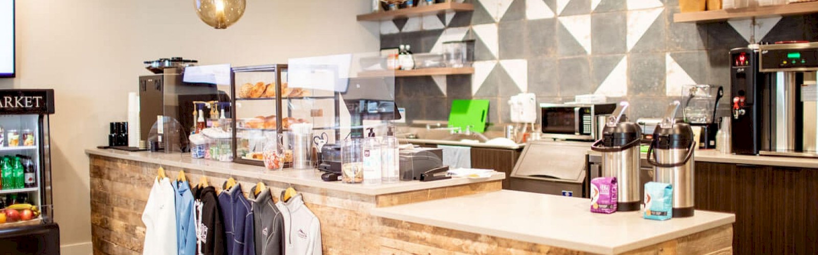 A modern coffee shop with a wooden counter, hanging lights, coffee machines, and merchandise displayed, including shirts and a small snack area.