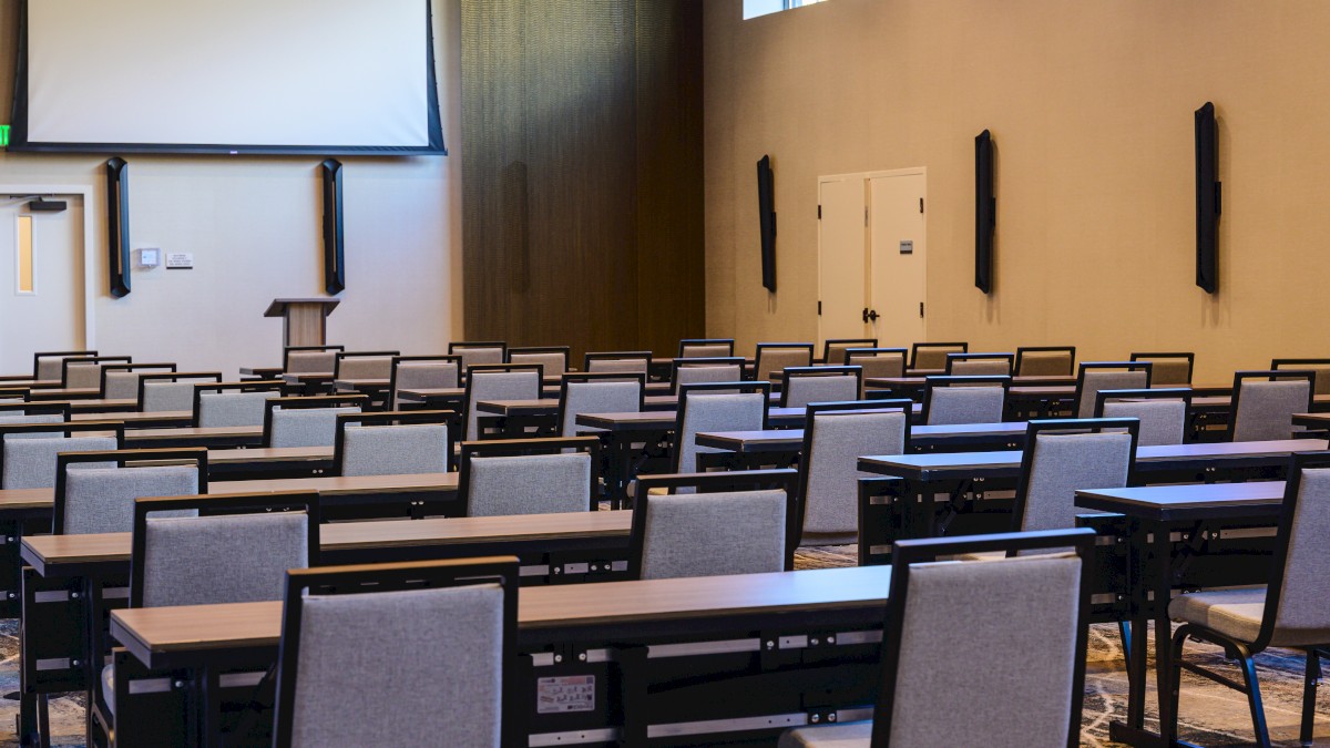 A large classroom or auditorium with rows of gray chairs and desks facing a stage, a projector screen, and a high ceiling with windows.