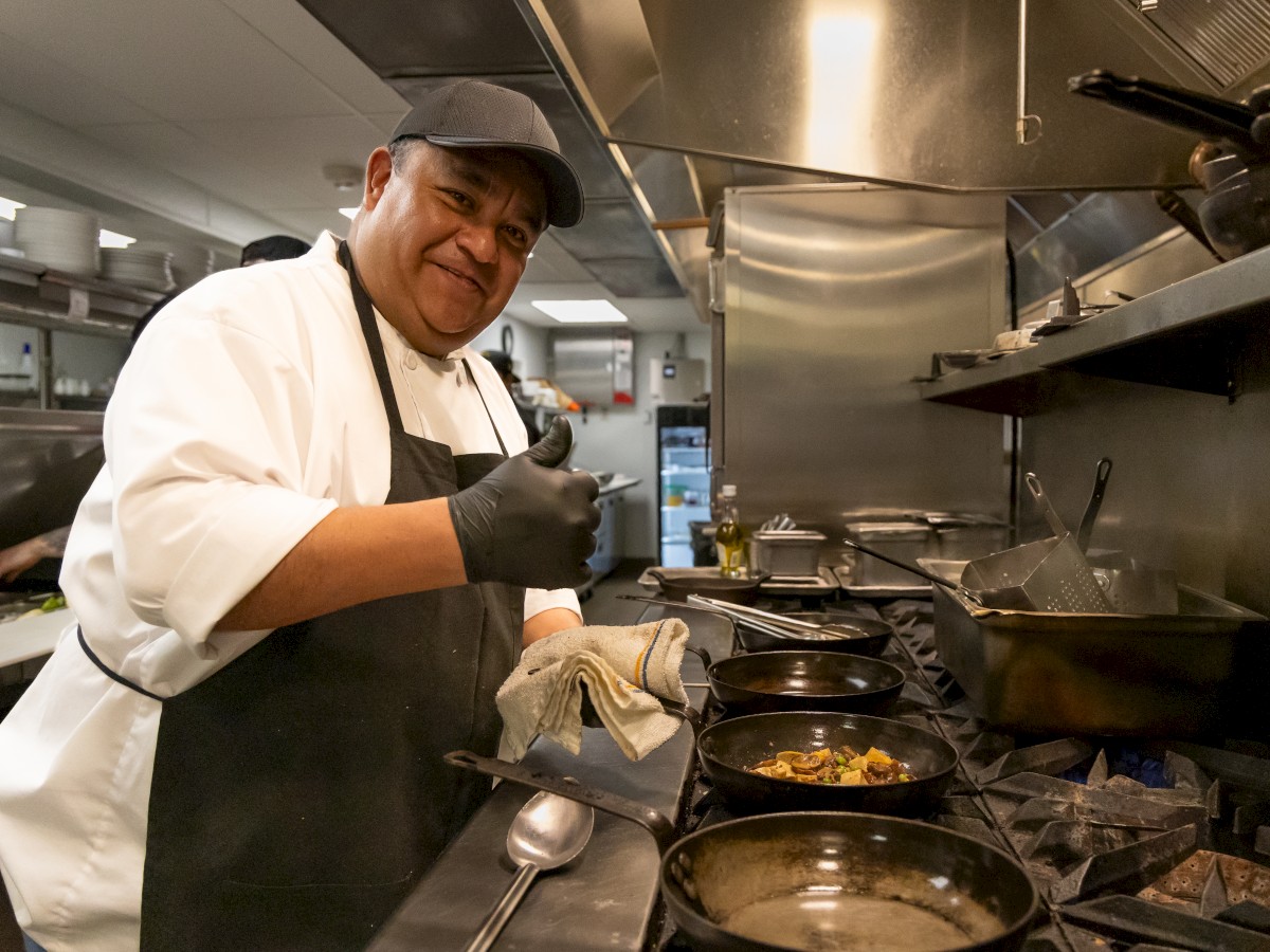 A chef in a kitchen gives a thumbs-up while cooking in multiple pans on the stove, wearing a white uniform, apron, and black cap.