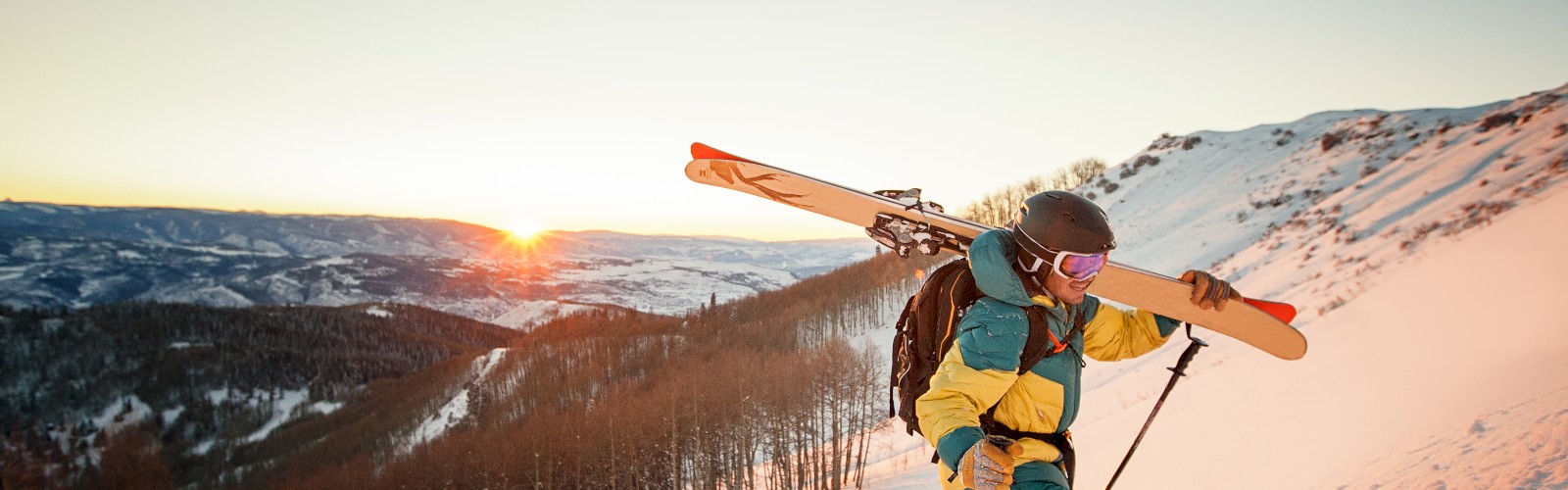 A skier carries skis up a snowy mountain slope at sunrise, with scenic forested hills and clear skies in the background.