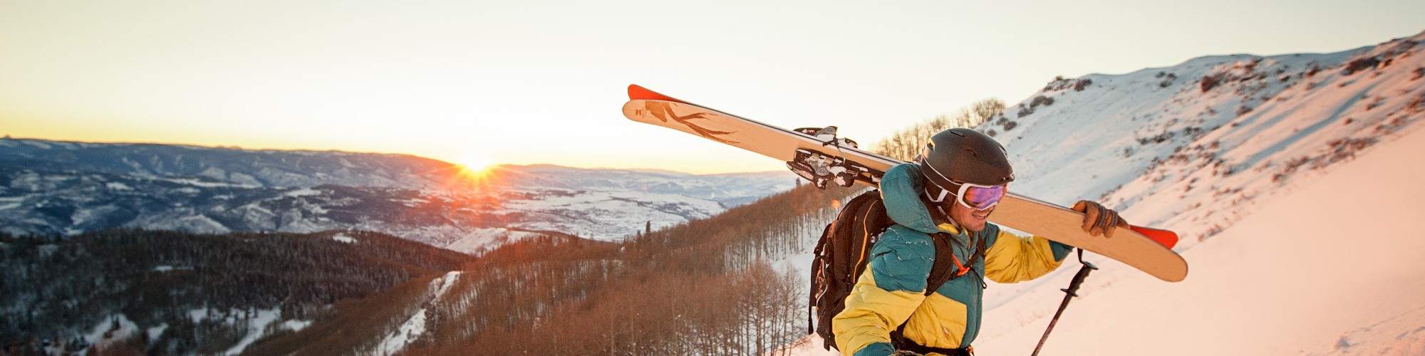 A skier carries skis up a snowy mountain slope at sunrise, with scenic forested hills and clear skies in the background.
