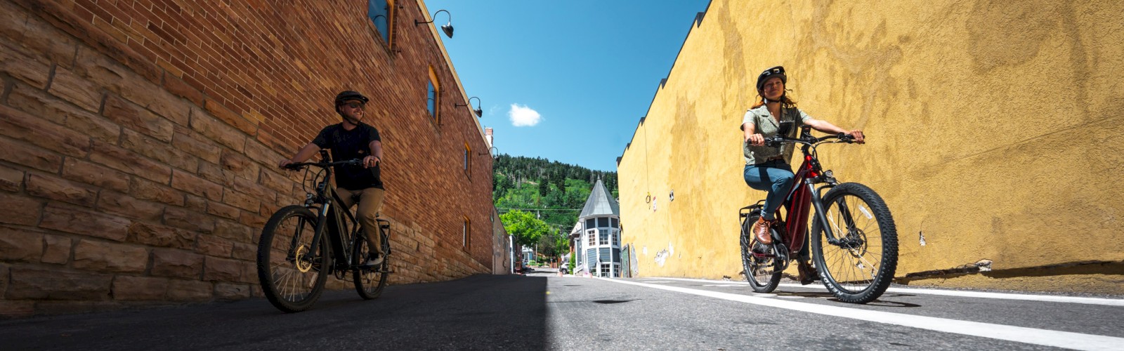 Two cyclists ride between two buildings on a narrow street with clear skies above and a distant green hill in the background.