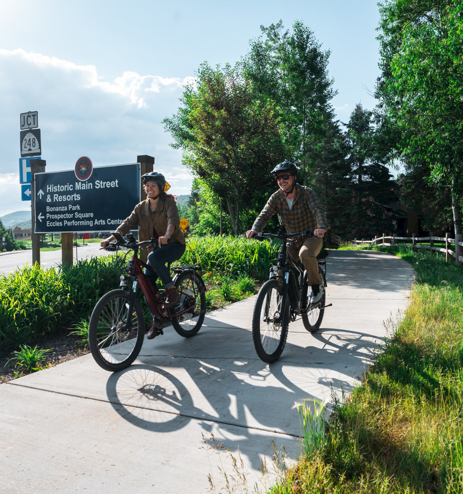 Two people are riding bicycles on a paved path next to a road. Greenery and trees surround the path, and a sign is visible in the background.