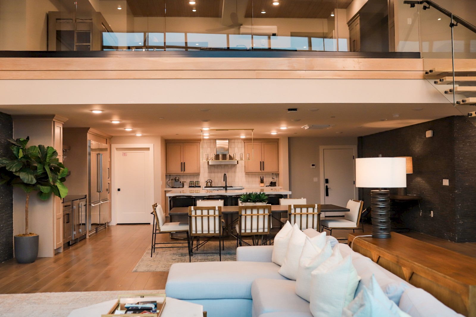 A modern open-plan living room and kitchen, featuring a sectional sofa, dining table, island, and stairs leading to an upper level.