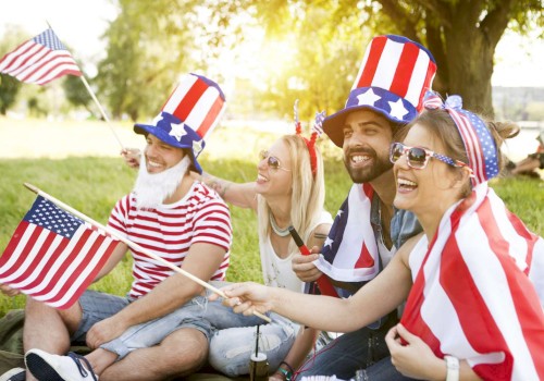 A group of people is sitting outdoors wearing red, white, and blue attire, holding American flags, and smiling, appearing to celebrate a patriotic event.