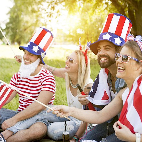 A group of people is sitting outdoors wearing red, white, and blue attire, holding American flags, and smiling, appearing to celebrate a patriotic event.