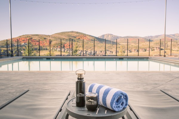A poolside scene with a striped towel, water bottle, and container on a small table, overlooking mountains through a glass fence.