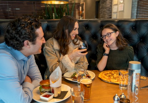 A family dines together at a restaurant, smiling and enjoying their meal, including burgers, pizza, and beverages.
