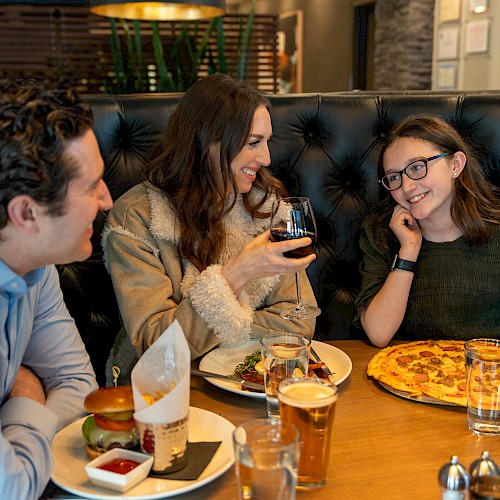 A family dines together at a restaurant, smiling and enjoying their meal, including burgers, pizza, and beverages.