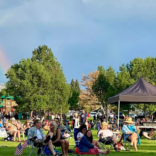 People are gathered in a park with lawn chairs, a canopy, and a rainbow in the sky visible in the background.