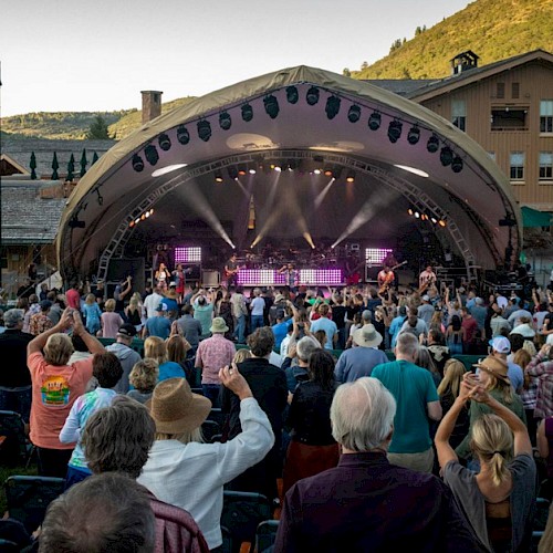 The image shows an outdoor concert with a large crowd facing a stage. Many people are standing and some have their hands raised, enjoying the event.