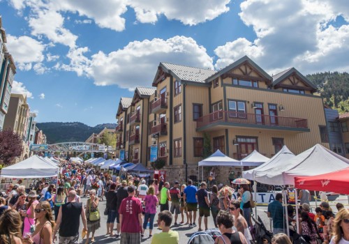 A street festival with many people attending, tents, and vendors. Residential buildings and mountains are seen in the background under a partly cloudy sky.