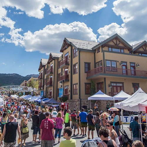 A street festival with many people attending, tents, and vendors. Residential buildings and mountains are seen in the background under a partly cloudy sky.